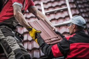 Two workers on a roof handing each other roofing materials.